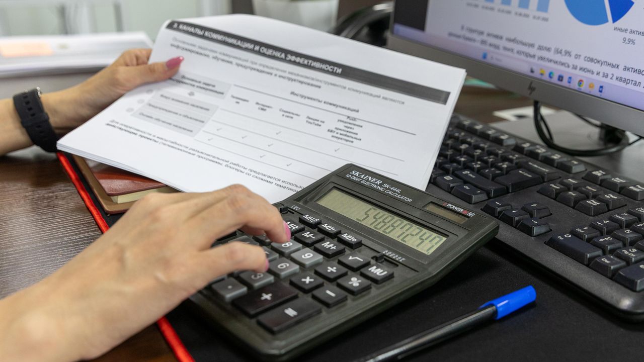 Una persona revisa un documento financiero con una calculadora en un escritorio, con un gráfico circular visible en la pantalla del ordenador detrás